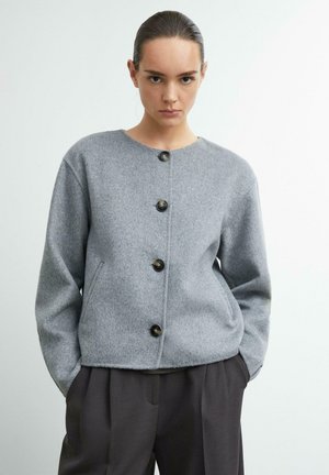 Frau trägt eine graue, mit Knöpfen versehene Wolljacke mit Taschen und dunkelgraue Hose, steht mit den Händen in den Taschen vor einem einfarbigen Hintergrund.