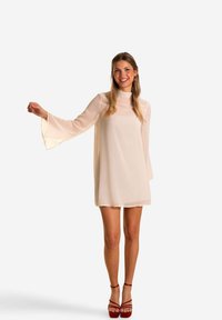 Robe courte en crème avec un col haut et des manches évasées, fabriquée à partir d'un tissu transparent. Portée avec des sandales à talons rouges.