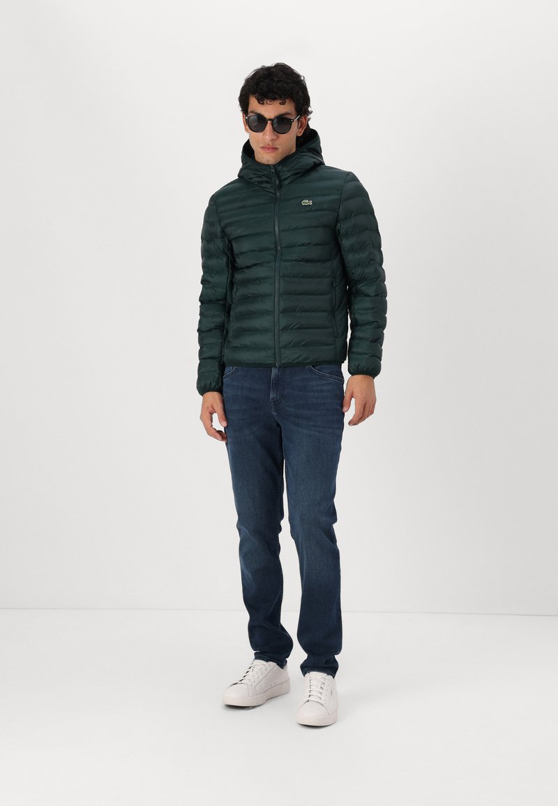 Chaqueta acolchada verde con diseño de costuras horizontales, cierre delantero con cremallera y capucha, combinada con vaqueros oscuros y zapatillas blancas.