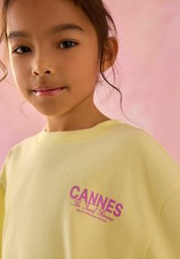Žuta pamučna majica s ružičastim grafikonom koji sadrži natpise "CANNES" i "Francuska rivijera". Okrugli izrez i širok kroj.