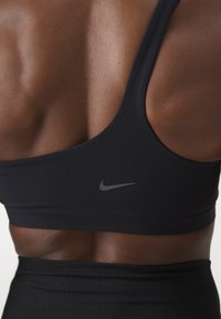 Svart sportsbh med jevn tekstur og brede stropper, med en grå Nike-logo på siden. Bakdesignet er minimalistisk og støttende.