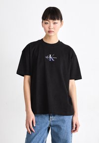 Crna pamučna majica s opuštenim krojem, rebrastim okruglim izrezom i središnjim grafičkim logom u ljubičastoj i bijeloj boji koji glasi "Calvin Klein Jeans".