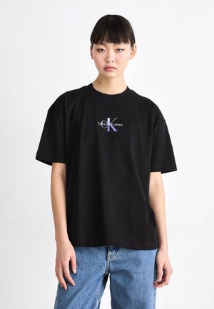 Juoda medvilninė marškinėliai su laisvu kirpimu, grublėto apvalaus kaklo ir centruotu grafikiniu logotipu purpurine ir balta spalvomis, skaitant "Calvin Klein Jeans."