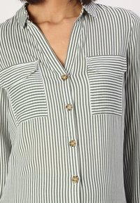 Blusa a righe verdi e bianche con scollatura a V, due taschini sul petto e chiusura con bottoni, realizzata in tessuto leggero. Presenta un orlo arrotondato e bottoni beige.