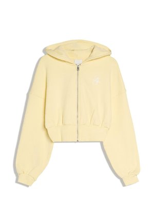 Svetložltá krátka mikina s kapucňou, predným zipsom, dlhými balónovými rukávmi, elastickými manžetami a malým bielym vyšívaným logom na hrudi.