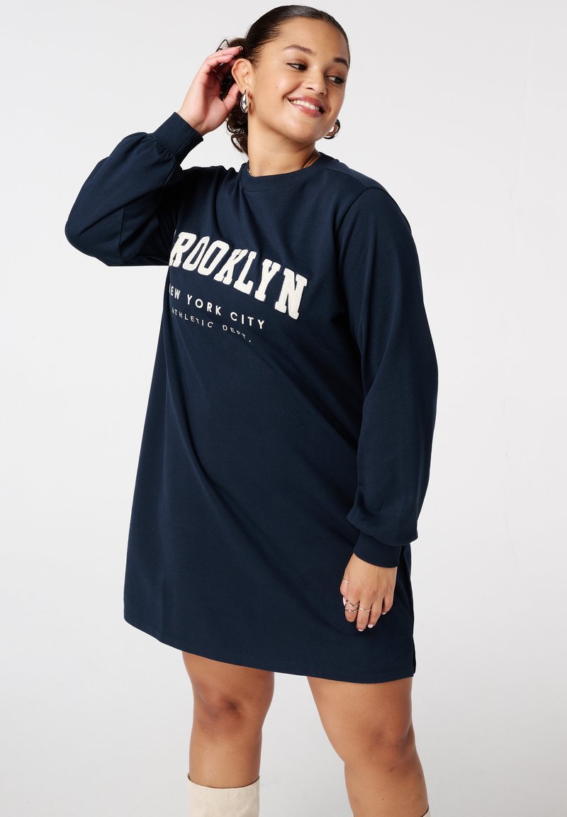 Robe-sweat bleu marine à manches longues, arborant "BROOKLYN NEW YORK CITY" en lettres blanches sur la poitrine. Tissu doux, coupe confortable.