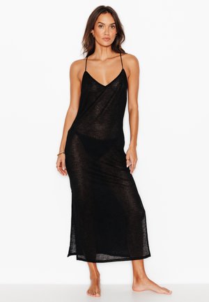 Vrouw draagt een lange zwarte doorschijnende slipjurk met dunne bandjes, staat blootsvoets tegen een witte achtergrond.