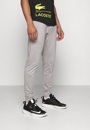 Homme portant un pantalon de survêtement gris, des baskets Nike noires et blanches, et un t-shirt Lacoste noir avec un logo jaune, debout sur un sol blanc uni.