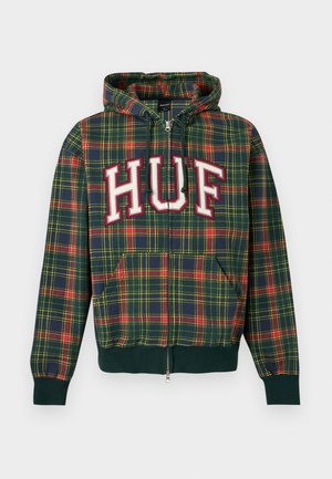 Jeune homme portant un sweat à capuche zippé à carreaux verts, rouges et jaunes avec l’inscription « HUF » sur la poitrine, assorti à un pantalon noir.
