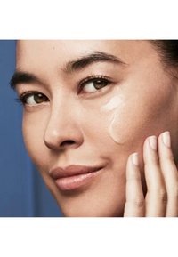 Produit de soins de la peau appliqué sur un teint clair et lisse. Gros plan montrant une petite quantité de crème sur la joue et des doigts touchant légèrement le visage.
