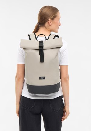 Mujer vistiendo una camiseta blanca y jeans negros, con una mochila beige y negra con cierre de hebilla en la parte superior, vista desde la espalda.