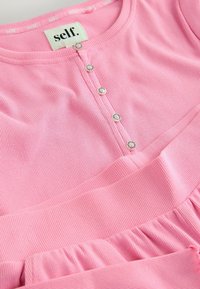 Top in tessuto a coste rosa con scollatura rotonda, caratterizzato da bottoni decorativi e un orlo arricciato. L'etichetta riporta la scritta "self." in bianco.