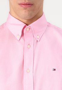 Lähikuva henkilöstä, joka käyttää vaaleanpunaista nappikauluksista paitaa, jossa on pieni suorakaiteen muotoinen lipun logo rinnassa.