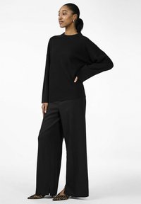 Maglione nero oversize con maniche lunghe, abbinato a pantaloni neri a gamba larga. Gli accessori includono scarpe con stampa leopardata e orecchini.