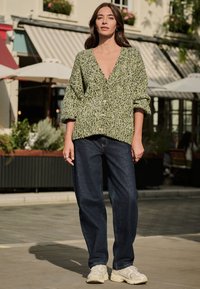 Grøn strikket cardigan med store ærmer, V-udskæring og knapper, kombineret med løse mørke denimjeans og beige sneakers.