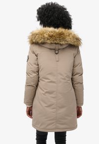 Cappotto trapuntato beige con cappuccio in pelliccia sintetica, chiusura con zip e vita regolabile. Presenta una patch con logo sulla manica e un design di lunghezza media.