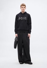 Μαύρο φούτερ με κουκούλα με το λογότυπο "BOSS" σε γκρι, συνδυασμένο με φαρδιά μαύρη παντελόνια. Κρατάει μια μαύρη δερμάτινη τσάντα. Το λουκ ολοκληρώνουν τα φλατ παπούτσια.