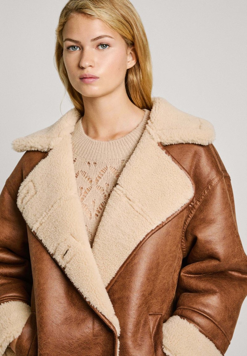 Manteau en cuir marron avec un col et une doublure en polaire beige. Il présente un large revers et de profondes poches. Superposé sur un haut en tricot beige.