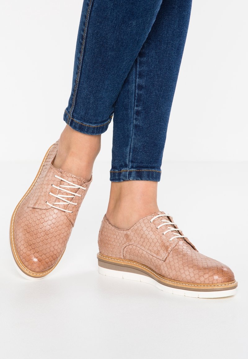 Pieds chaussés de chaussures à lacets texturées marron clair associées à un jean bleu foncé retroussé sur un fond blanc.