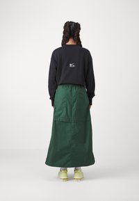 Person med flätat hår bär en svart Nike-huvtröja, en mörkgrön lång kjol och ljusgula Nike-sneakers, stående vänd bort.