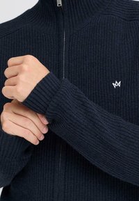 Námořnická modrá žebrovaná pletená bunda na zip s vysokým límcem, texturovanou látkou a malým bílým vyšitým logem na prsou.