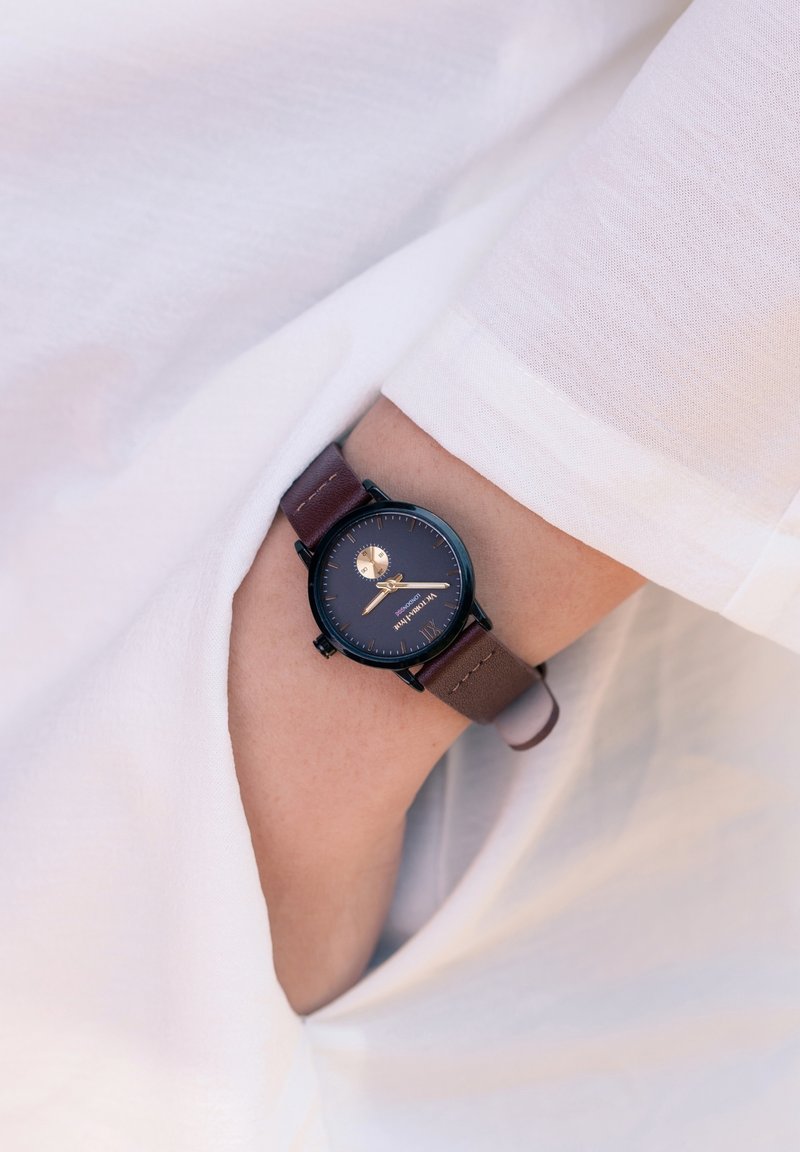Poignet avec une montre à cadran noir et bracelet en cuir marron partiellement glissée dans le poignet et la poche d'une chemise blanche à manches longues.