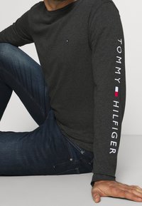 Mężczyzna siedzący w pozycji skrzyżowanej, ubrany w ciemnoszary sweatshirt Tommy Hilfiger z logo na klatce piersiowej i rękawie, w połączeniu z ciemnoniebieskimi dżinsami.