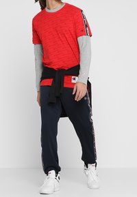 Rød grafisk t-shirt lagdelt over en grå langærmet top, parret med navy sweatpants med sidelister og hvide sneakers.