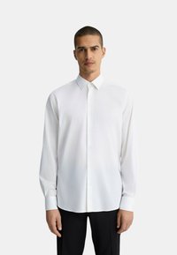 Camicia bianca a maniche lunghe con colletto e chiusura a bottoni, realizzata in tessuto liscio, indossata con pantaloni neri.
