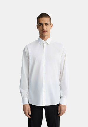 Camicia bianca a maniche lunghe con colletto e chiusura a bottoni, realizzata in tessuto liscio, indossata con pantaloni neri.