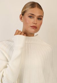 Weißer gerippter Pullover mit hohem Kragen und langen Ärmeln, der eine weiche Textur und subtile Variationen im Strickmuster zeigt.