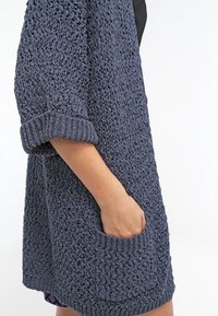 Öppen stickad cardigan i mörkblått med uppvikt ärm och en framficka. Tillverkad av texturerad garn, med en lös och avslappnad passform.