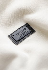 Étiquette noire rectangulaire « ombre menswear » cousue sur un tissu lisse beige clair avec des plis subtils.