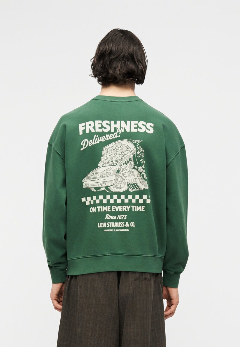 Personne de dos portant un sweat-shirt vert avec un motif de voiture et une tranche de pizza, le texte indique « Fraîcheur livrée ! À l'heure à chaque fois. »