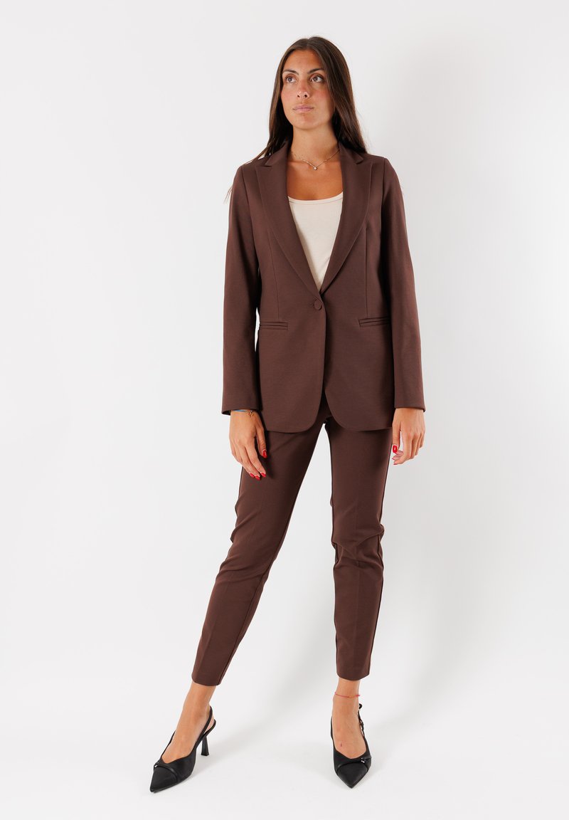 Donna in piedi che indossa un completo marrone con giacca aderente e pantaloni, una maglia panna e décolleté neri a punta su uno sfondo bianco.