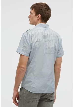 Mann im Profil, der ein hellblaues kurzärmeliges Hemd mit blassem weißen Text auf dem Rücken und graue Hosen trägt.