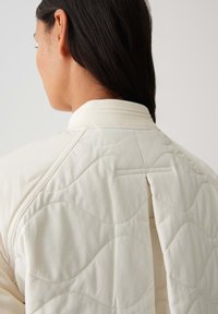 Veste en crème matelassée avec un col haut et de subtils motifs en vagues à l'arrière. Comprend un détail de fente dans le dos pour un style et une fonctionnalité supplémentaires.
