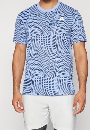 Male wearing a light blue, short-sleeve shirt with dark blue wavy dot patterns and white shorts, standing against a plain background.