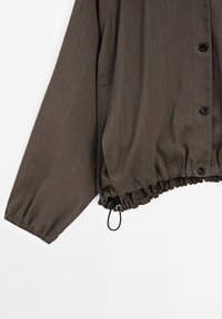 Giacca leggera marrone con chiusura a bottoni, polsini elasticizzati e orlo arricciato con cordino per un fit regolabile. Texture liscia.