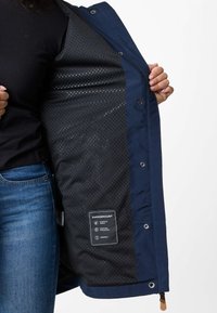 Personne tenant ouverte une veste bleu marine montrant sa doublure intérieure noire perforée avec une étiquette de marque Swedemount à l'intérieur.