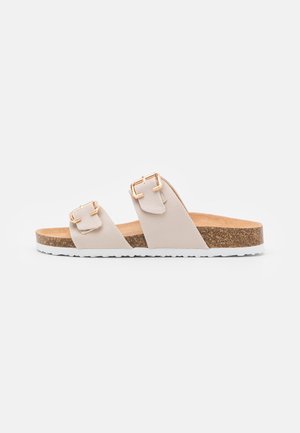 Beige læder slidesandal med to justerbare remme, gyldne spænder, korkfodtøj og hvid tekstureret sål.