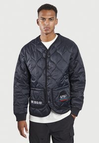 Chaqueta negra acolchada con un frente botonado, que presenta un parche de la NASA y texto bordado en blanco. Tejido texturizado con un acabado suave.