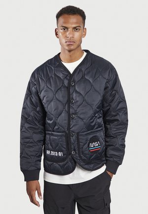Schwarze gesteppte Jacke mit einem geknöpften Frontverschluss, ausgestattet mit einem NASA-Patch und weißem, gesticktem Text. Strukturierter Stoff mit glatter Oberfläche.