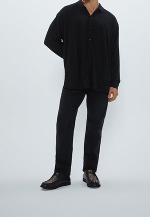 Chemise noire à boutons avec une coupe décontractée et une texture côtelée subtile, associée à un pantalon sombre et des chaussures noires avec un design à rayures dorées.