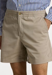 Béžové šortky s elastickým pasem, zapínáním na knoflík a bočními kapsami. Hladká tkanina a jemný detail loga na lemu.
