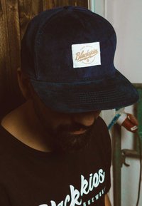 Cappello in corduroy blu navy con visiera piatta e un'etichetta bianca con il testo "Blackskies." Indossato da una persona con una maglietta nera.