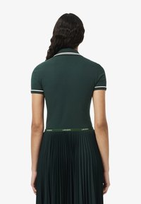 Polo shirt in donkergroen katoen met witte afwerking bij de kraag en mouwen; heeft een geplooide rok met een groene Lacoste tailleband.