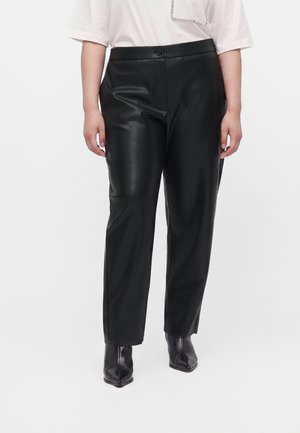 Persona che indossa pantaloni in pelle nera a gamba dritta, stivali neri con punta appuntita e una camicia bianca con dettaglio decorativo sul petto.