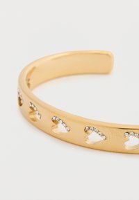 Χρυσό βραχιόλι μανσέτα διακοσμημένο με κοψίματα σε σχήμα καρδιάς, στολισμένα με μικρές στρογγυλές στρας κατά μήκος της ταινίας. Ομαλό μεταλλικό φινίρισμα.