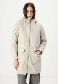 Beige hettejakke med glidelås foran, justerbar snor i midjen og to frontlommer; glatt tekstur og svakt blank finish.